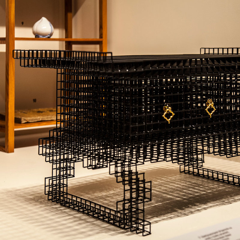 1. Widok wystawy. Koreański stolik do czytania wykonany z czarnego stalowego drutu o współczesnej, ażurowej formie. W tle drewniany regał.