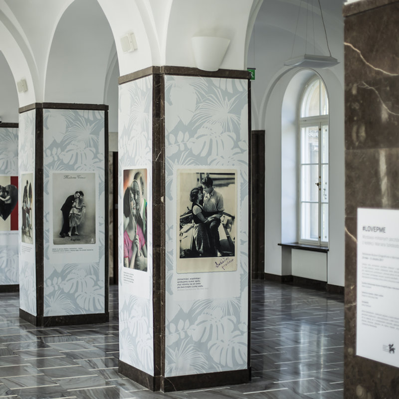 Widok wystawy z umieszczonymi na obudowie filarów przedrukami pocztówek z pierwszej połowy XX wieku, przedstawiających pary w romantycznych objęciach.