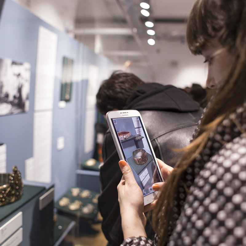 Zdjęcie przedstawia kobietę, zwiedzającą wystawę, która fotografuje telefonem komórkowym obiekty prezentowane na wystawie.