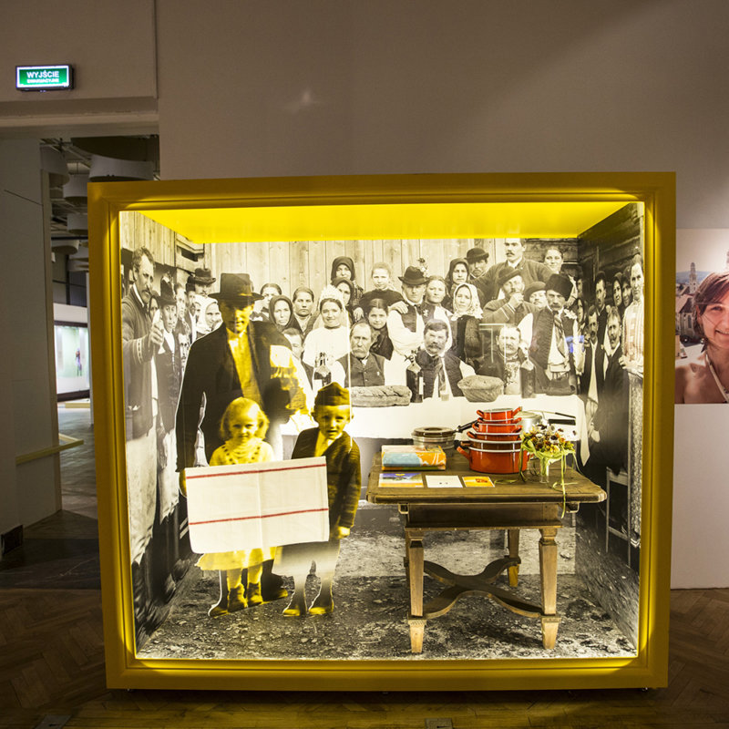 Żółto obramowana diorama, której tło stanowi czarno - białe zdjęcie uczty weselnej z początku XX w. Przed zdjęciem stoi drewniany stół a na nim stos pomarańczowych garnków, aluminiowy rondel oraz zasuszony bukiet weselny. Obok stołu stoją naturalnych rozmiarów, wycięte z fotografii, czarno-żółte figury mężczyzny i dwojga dzieci (chłopca i dziewczynki) idących na wesele.