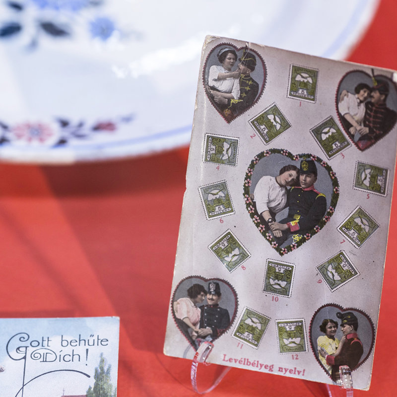 Miłosna kartka z 1916 roku. Na kartce w centralnej części oraz w rogach w ramkach w kształcie serca przedstawione są pary w romantycznych objęciach, pomiędzy nimi na całej kartce naklejone są zielone znaczki pocztowe. Na dole kartki węgierski napis, który można przetłumaczyć „Miłosny język znaczków pocztowych”.