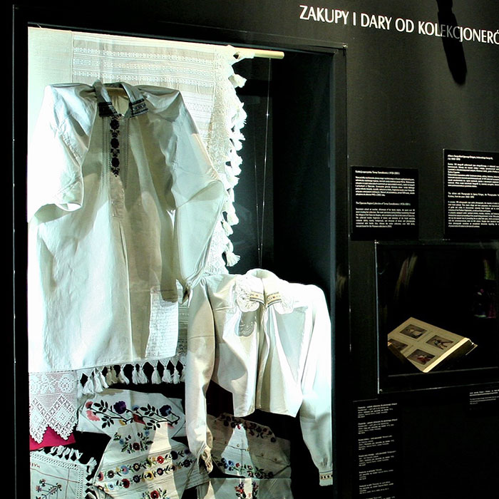 Dwie białe koszule z haftowanymi elementami wiszące w gablocie: jedna kobieca krótka, druga koszula pana młodego długa, poniżej haftowane elementy: powłoczka na poduszkę, ręcznik obrzędowy, chusteczka.