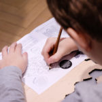 Fotografia kolorowa przedstawiająca dziecko z ołówkiem w ręku, rozwiązujące zadania na kartce papieru.