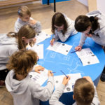 Fotografia kolorowa przedstawiająca dzieci siedzące wokół niebieskiego stolika, rozwiązujące zadania na kartkach papieru.