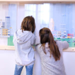 Fotografia kolorowa przedstawiajaca dwie dziewczynki przyglądające się obiektom za szklaną gablotą. W gablocie: wizerunki bloków i miejskiego krajobrazu.
