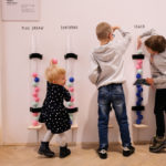 Fotografia kolorowa przedstawiająca trójkę dzieci w różnym wieku. Dzieci wrzucajć kolorowe, plastikowe kulki do przezroczystych, walcowatych pojemników.