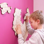 Fotografia kolorowa przedtawiająca chłopca stojącego pod różową ścianą, dopasowującego fragmenty układanki mapy umieszczone na ścianie.