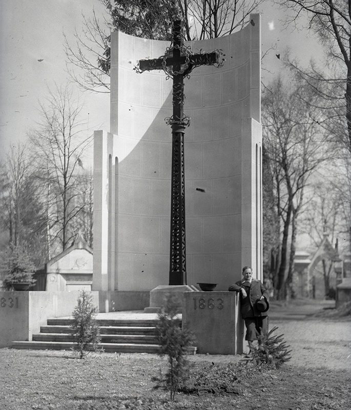 Krzyż powstańczy z cmentarza komunalnego w Nowym Sączu_Arch. PME N.4053_1. Wysoki, ażurowy, czarny krzyż otoczony białym półkolistym murem. Prowadzą do niego trzy stopnie schodów. Jest umieszczony na podniesieniu z wysuniętymi postumentami z napisami: z lewej 1831, z prawej 1861. Z prawej strony krzyża stoi wsparty o postument mężczyzna.