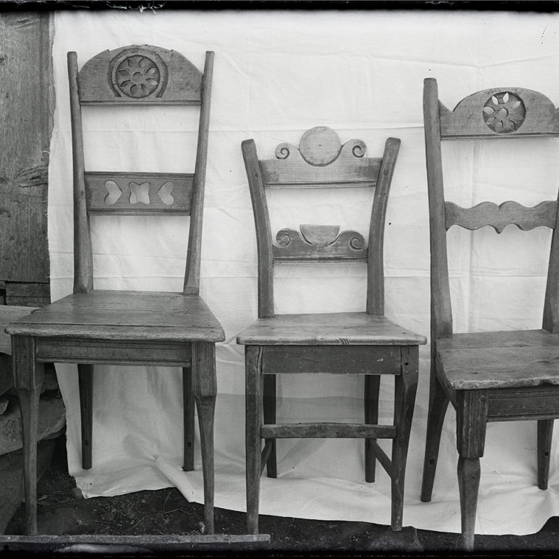 Meblarstwo ludowe. Krzesła ozdobne_Arch. PME N.4074_1. Trzy krzesła, różnych wielkości, z rzeźbionymi oparciami, na tle białej tkaniny. Z lewej strony fragment drzwi.