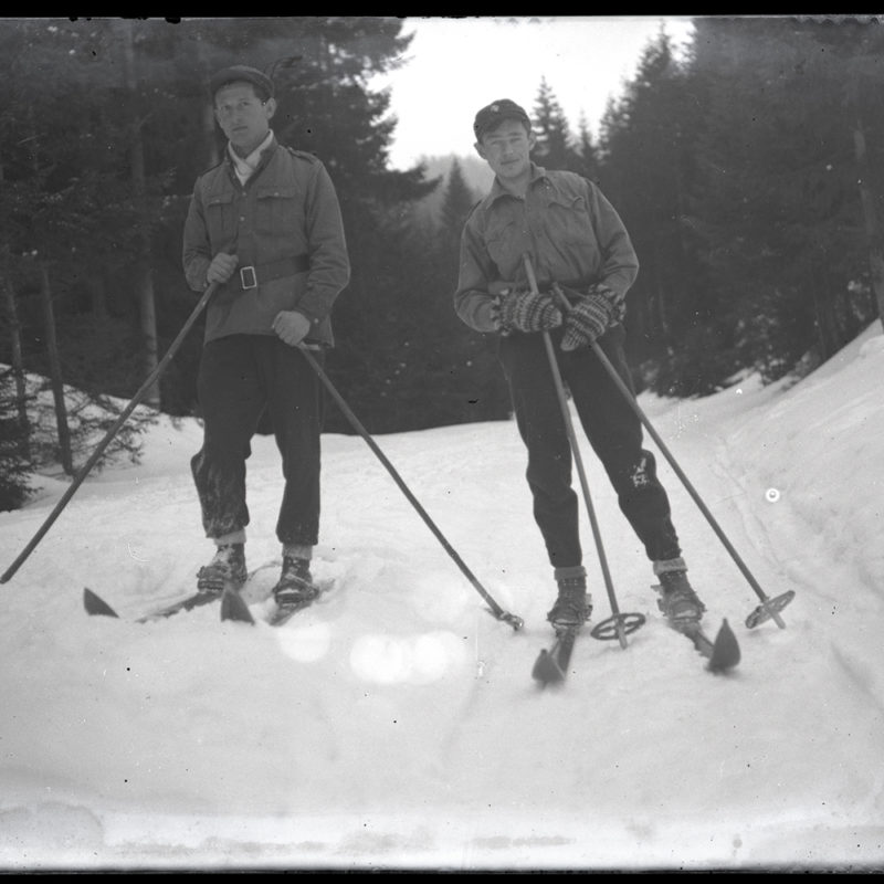 Mieczysław Cholewa na nartach z kolegą ok. 1935 r. _Arch. PME N.4125_1. Na górskiej, zasypanej śniegiem drodze, dwaj młodzi mężczyźni z nartami przypiętymi do butów oraz kijkami w rękach. Pozują do zdjęcia.