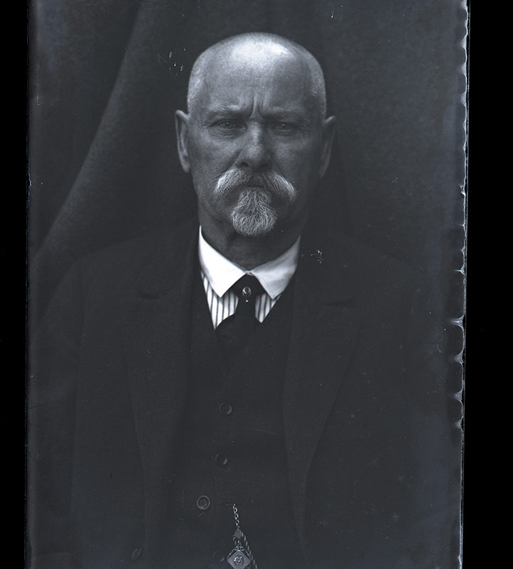 Portret starszego mężczyzny (ojciec Mieczesława Cholewy)_Arch. PME N.4127_4. Portret starszego mężczyzny na tle kotary, w ujęciu frontalnym, do pasa. Ubrany jest w rozpiętą czarną marynarkę, pod nią nosi ciemną kamizelkę zapinaną na małe guziki oraz koszulę z białym kołnierzykiem i ciemny krawat.