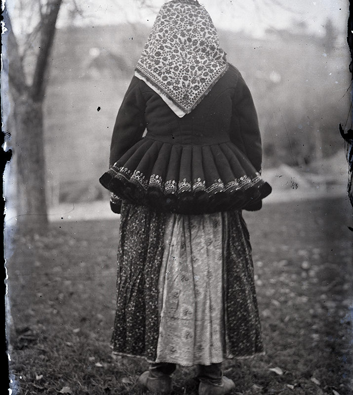 Strój. Kobieta w stroju Lachów Sądeckich_Arch. PME N. 4148_1. Stojąca tyłem kobieta w spódnicy w kwiaty, w kurtce z baskinką haftowaną u dołu i chustą w kwiatowy wzór na głowie - strój Lachów Sądeckich.