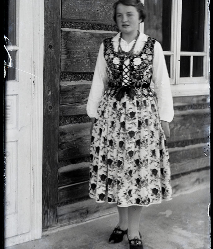 Strój. Kobieta w stroju łąckim z Kamienicy_Arch. PME N.4146_3. Kobieta w spódnicy w kwiaty, białej bluzce i bogato zdobionym gorsecie na tle drewnianego domu - strój łącki z Kamienicy