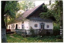 Kolorowe zdjęcie całego zalipiańskiego domu pośród zieleni: traw, krzewów i drzew. Na dachu domu położona strzecha, wszystkie ściany pomalowano na biało i bogato ozdobiono charakterystycznymi motywami kwiatowymi.