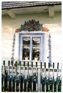 Kolorowe zdjęcie fragmentu zalipiańskiego domu - w centrum zdjęcia znajduje się pomalowane na niebiesko okno. Na białym domu wokół okna wymalowane zostały charakterystyczne dla Zalipia kwiaty. Przed domem skromny, drewniany, biały płotek