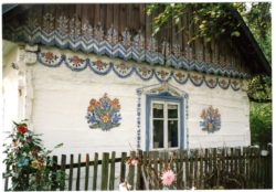 Kolorowe zdjęcie fragmentu zalipiańskiego domu - w centrum zdjęcia znajduje się pomalowane na niebiesko okno. Na białym domu wokół okna wymalowane zostały charakterystyczne dla Zalipia kwiaty. Przed domem skromny, drewniany płotek