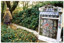 Zdjęcie kolorowe, na którym po prawej stronie znajduje się wejście do ziemianki znajdującej się pośród zieleni wymalowanej w zalipiańskie kwiaty. Na ziemiance data 1944 rok. Po lewej stronie starsza uśmiechnięta kobieta malująca wzór na murku.