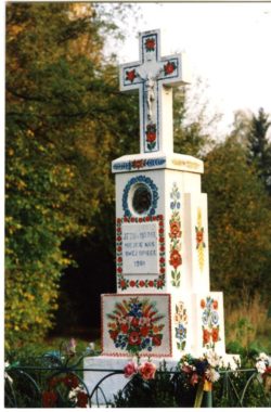 Kapliczka malowana w charakterystyczne zalipiańskie kwiaty na białym tle. na wysokim podeście stoi murowany krzyż.