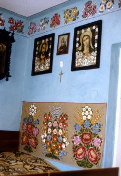 Wnętrze zalipiańskiego domu. Miejsce przy łóżku, ściany pomalowane na niebiesko i udekorowane. U dołu ściany wisi makatka z namalowanymi kolorowymi kwiatami, powyżej zbiór religijnych obrazków oraz mały krzyż.