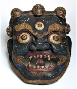 Zdjęcie kolorowe przedstawiające tybetańską maskę w kolorze ciemnogranatowym z wizerunkiem twarzy o wyszczerzonych groźnie kłach. Widoczne dwie wybałuszone i przekrwione gałki oczne oraz trzecia, mniejsza na środku czoła. Nad czołem wizerunek trzech czaszek. Wokół oczu i ust widoczne zdobienia w kolorze złotym.
