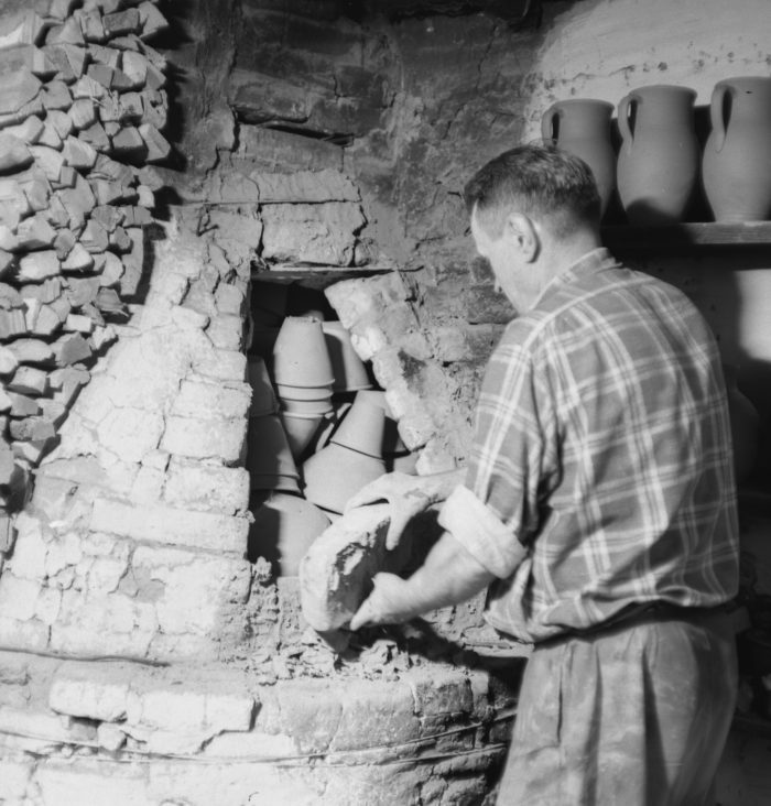 Czarnobiałe zdjęcia przedstawiające garncarza z Siemiatycz, który zamyka piec, by wypalić naczynia. Gliniane przedmioty ściśle układają się w kamiennym piecu opalanym drewnem.