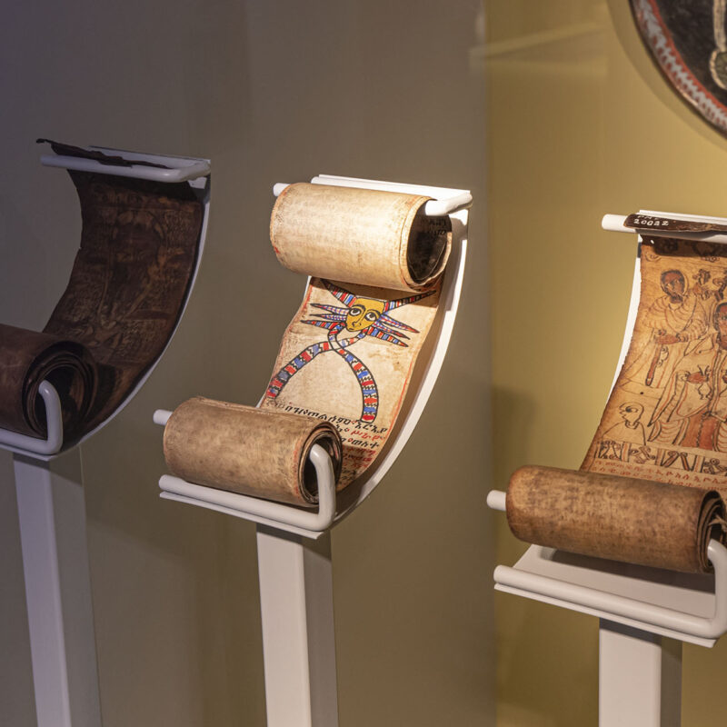 Widok wystawy. Na trzech białych stojakach z uchwytami prezentowane są trzy etiopskie zwoje magiczne, czyli zwijane w rulon rękopisy z ilustracjami.