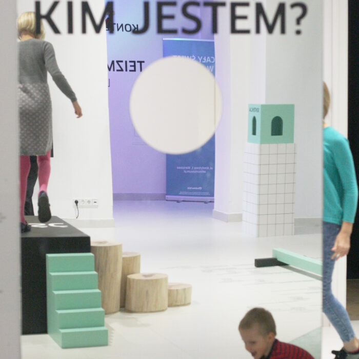 Zdjęcie kolorowe przedstawia lustro z naklejonym u góry pytaniem "Kim jestem?". Po środku lustra wycięta została okrągła dziura wielkości głowy. Pozostałą część fotografii buduje odbicie w lustrze, w którym widzimy dorosłą postać stojącą na podeście oraz dziecko bawiące się na podłodze.