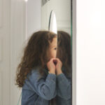 Zdjęcie kolorowe przedstawia dziewczynkę w wieku około 5 lat, która zagląda przez wyciętą w lustrze dziurę.