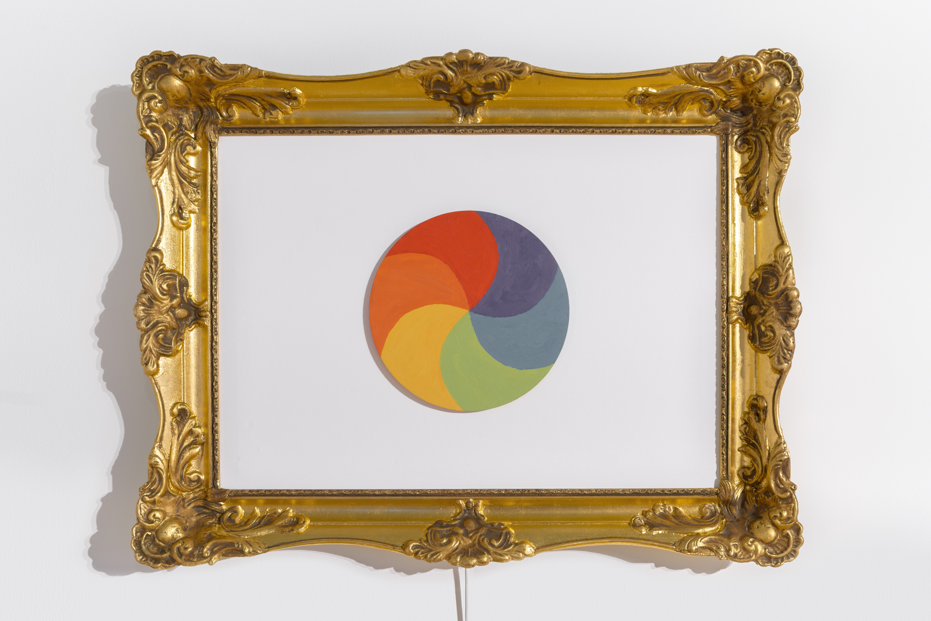 Złota, bardzo bogato zdobiona rama obrazu, , wewnątrz białe tło. Na środku tła koło pomalowane w sześciu kolorach: ciemnoniebieskim, niebieskim, zielonym, żółtym, pomarańczowym i czerwonym. Fot Maciej Jędrzejewski