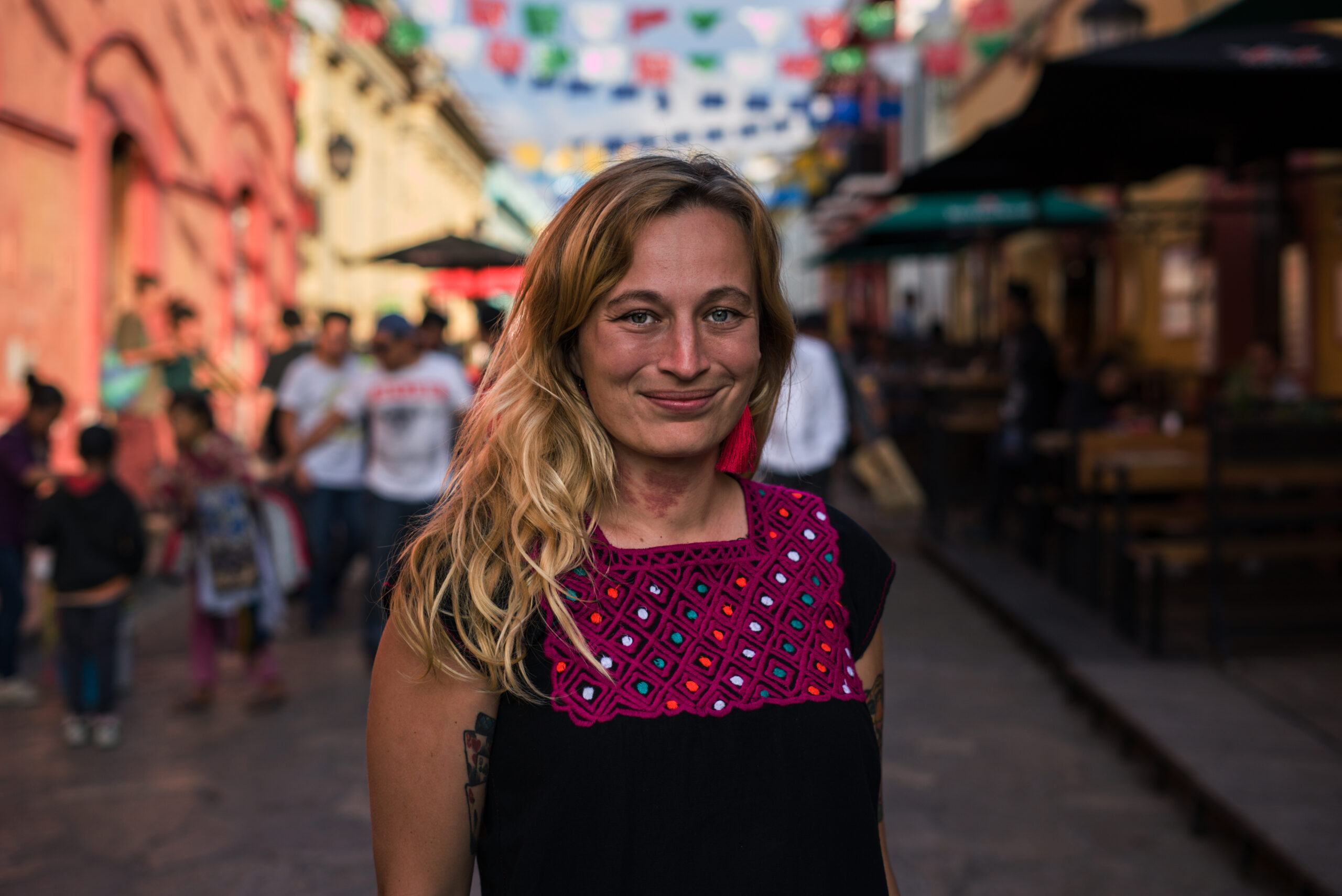Ola Synowiec na pierwszym planie, na tle ulicy w Meksyksu. Kobieta uśmiechnięta, rozpuszczone długie blond włosy, ubrana w bluzkę z dekoracją w stylu meksykańskim.