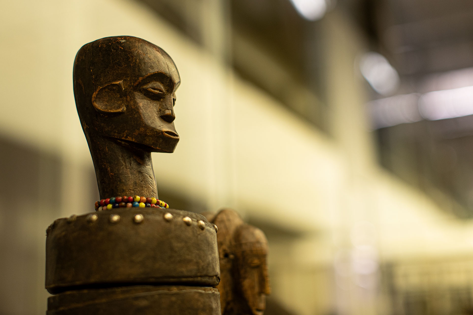 Zdjęcie przestawiające rzeżbę z wystawy "Afrykańskie wyprawy, azjatyckie drogi". Popiersie, głowa ukazana bokiem, postać bez włosów, koraliki dekoracyjne na szyii. 