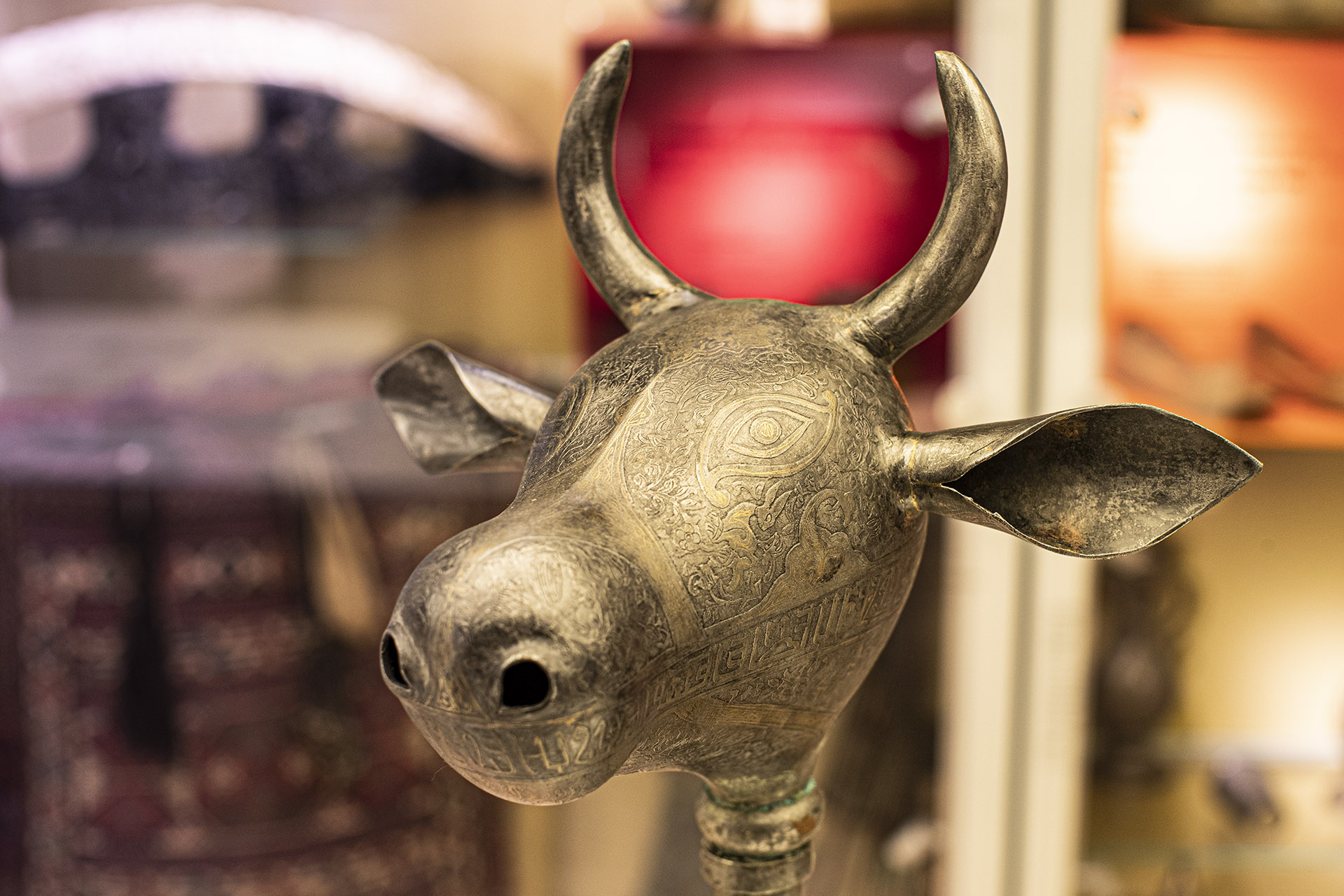 Zdjęcie przedstawia buławę o byczej głowie. Przedmiot wykonany z metalu. Głowa byk zdobiona ornamentem.