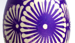 Zdjęcie kolorowe przedstawiające pisankę wielkanocną tworzoną metodą batikową. Jajko barwione na fioletowo z ozdobnymi okręgami imitującymi kwiaty znajduje się na białym tle.