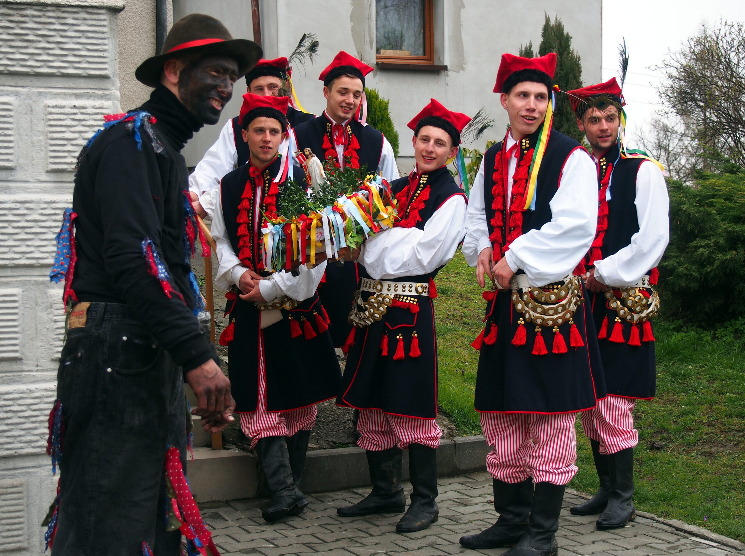 Uczestnicy obrzędu Siuda Baba przd domem. Sześciu mężczyzn ubranych w regionaln stroje krakowskie, jeden z usmoloną twarzą jako Siuda Baba.