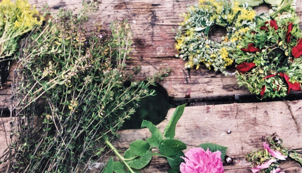 Zdjęcie przedstawia elementy potrzebne do robienia wianków. Na drewnianym taborecie ułożone kwiaty, zioła i gotowe wianki.