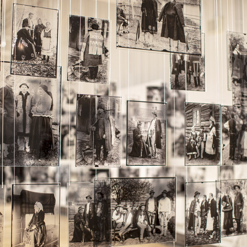 Widok na kilkadziesiąt powiększeń fotografii Mieczysława Cholewy wykonanych na płytkach plexi imitujących technikę negatywu szklanego, płytki różnych rozmiarów zawieszone są na żyłkach.