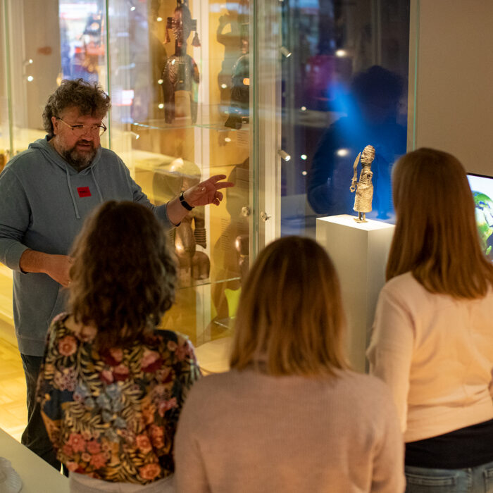 Dariusz Skonieczko podczas oprowadzania po wystawie Afrykańskie wyprawy, azjatyckie drogi wskazuje na niewielką figurkę wykonaną z metalu. Słuchają go trzy osoby.