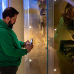 Na zdjęciu mężczyzna zwiedzający wystawę, fotografuje gablotę z obiektami na wystawie.
