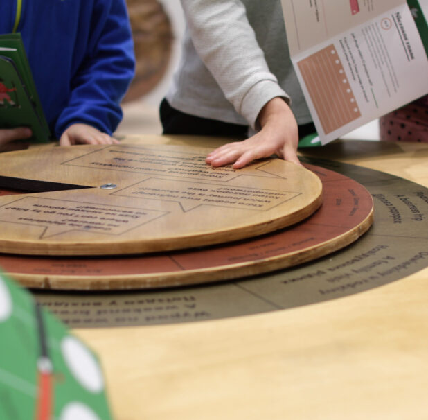 Zdjęcie z wystawy. Na stole okrągła plansza wykonana z drewna. Wokół stołu zgromadzona grupa dzieci. Na zdjęciu widać ich ręce, w których trzymają kolorowe kartki z zadaniami. Jedno z dzieci kręci okrągłą planszą.