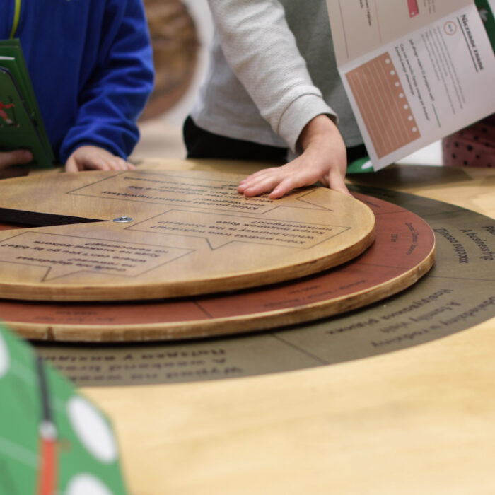 Zdjęcie z wystawy. Na stole okrągła plansza wykonana z drewna. Wokół stołu zgromadzona grupa dzieci. Na zdjęciu widać ich ręce, w których trzymają kolorowe kartki z zadaniami. Jedno z dzieci kręci okrągłą planszą.