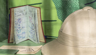 Na zdjęciu fragment wystawy. Na niejednorodnym zielonym tle ułożone przedmioty. Po lewej dwie książeczki. Jedna leży zamknięta na pierwszym planie. Za nią druga, otwarta, ustawiona pionowo, z pieczątkami na stronach. Po prawej jasnozielony kapelusz podróżnika