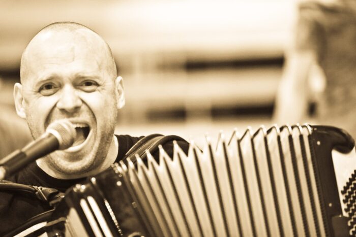 Portret Adama Struga wykonany podczas śpiewu; przed muzykiem znaduje się mikrofon oraz akordeon.
