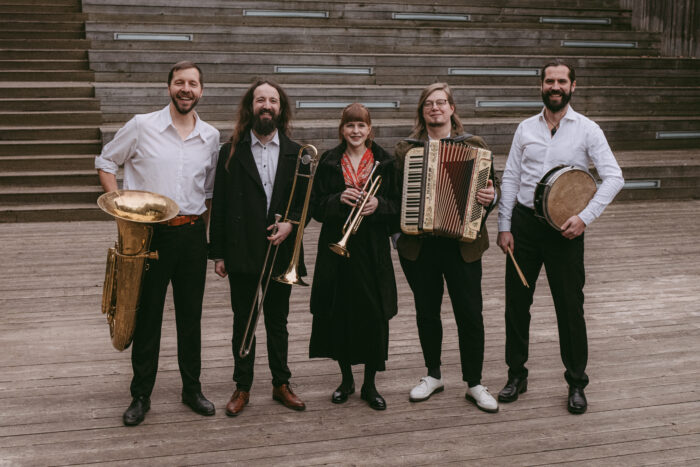 Zdjęcie przedstawiające skład grupy. Pięć osób ustawionych z rzędzie, trzymających instrumenty muzyczne. Znajdują się na drewnianej scenie.