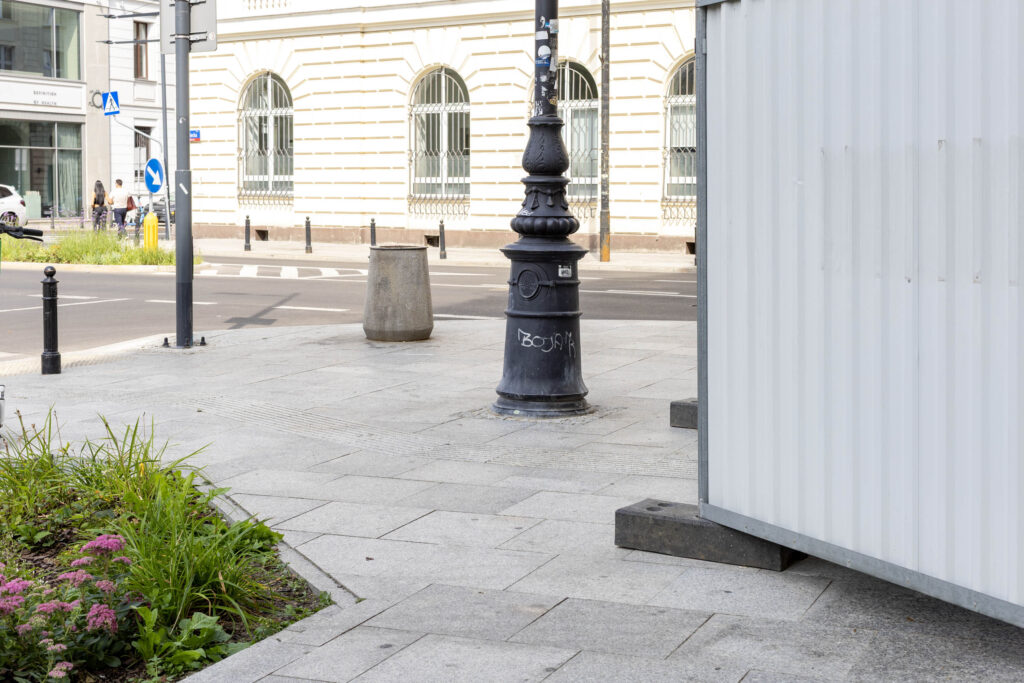 Chodnik przed muzeum ze ścieżką dotykową dla osób z niepełnosprawnością wzroku, częściowo zwężony przez białe metalowe ogrodzenie remontowe.